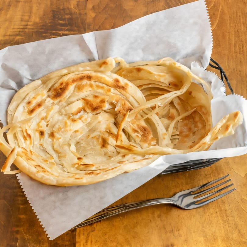 Flaky, golden-brown South Indian parotta bread in a basket with a fork on a wooden table.