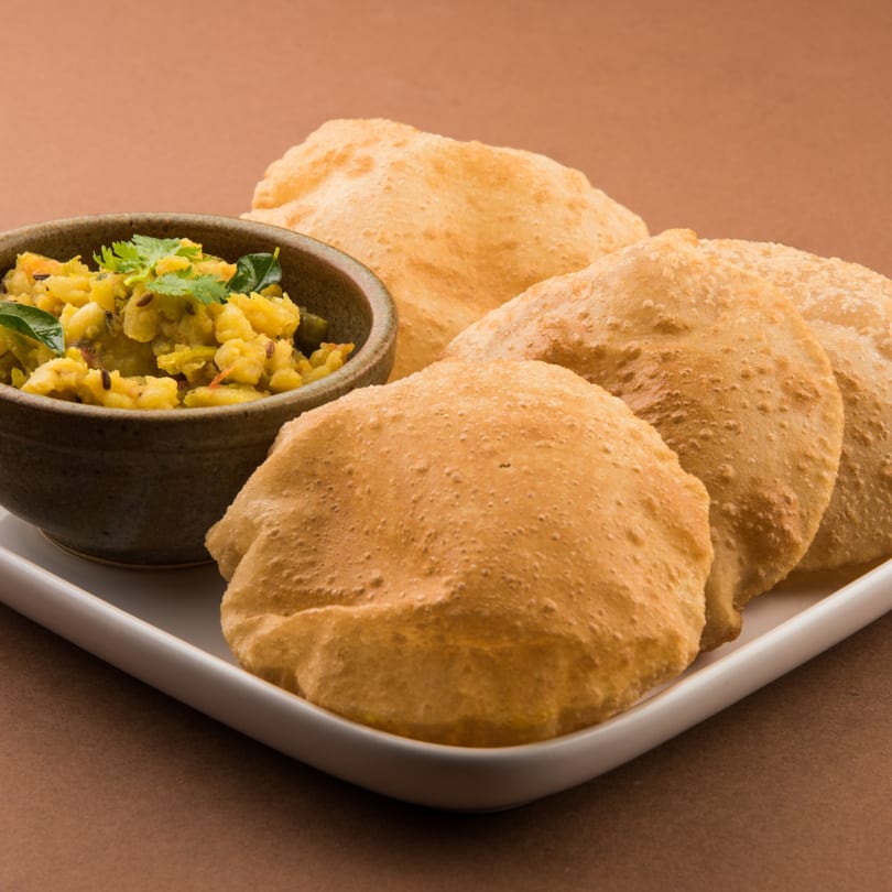 A bowl of potato curry with cilantro and cumin, served with several golden-brown puri on a white plate.