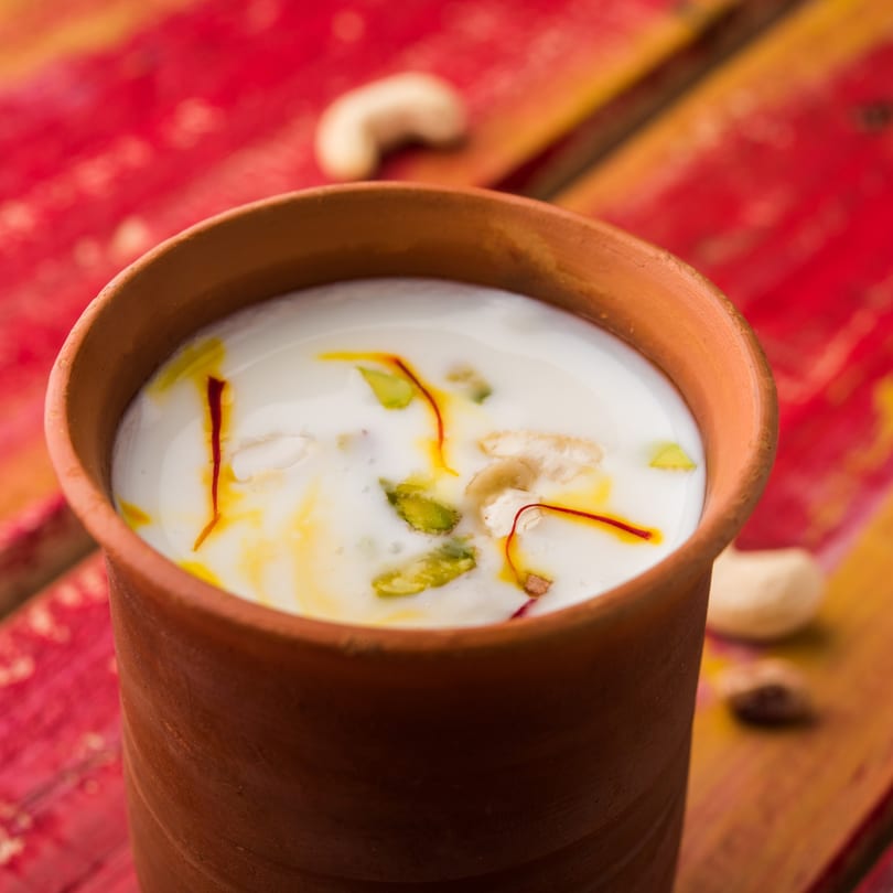 Sweet Lassi in a clay cup, garnished with saffron, pistachios, and cashews on a red and yellow surface.